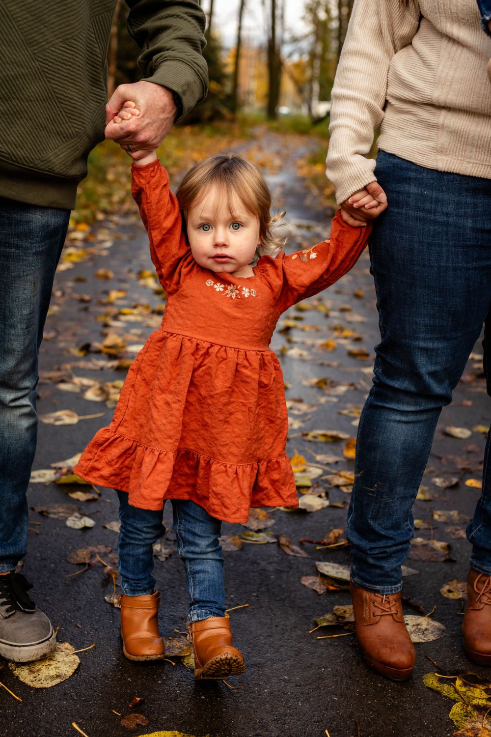 Small child in orange dress walking while holding hands on leaf-covered path.