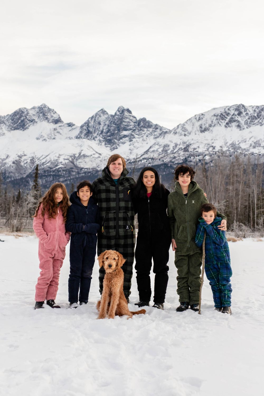 Family and dog pose together in snow with majestic mountain range backdrop.