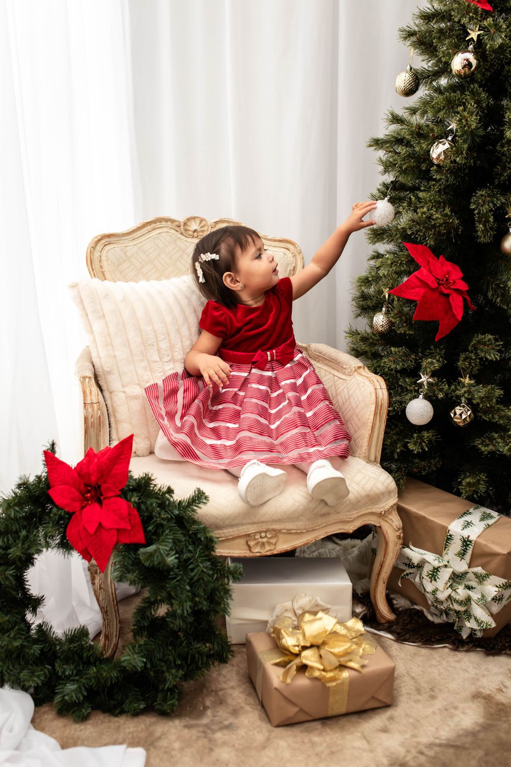 Small child in red and white dress reaching for ornament on Christmas tree.
