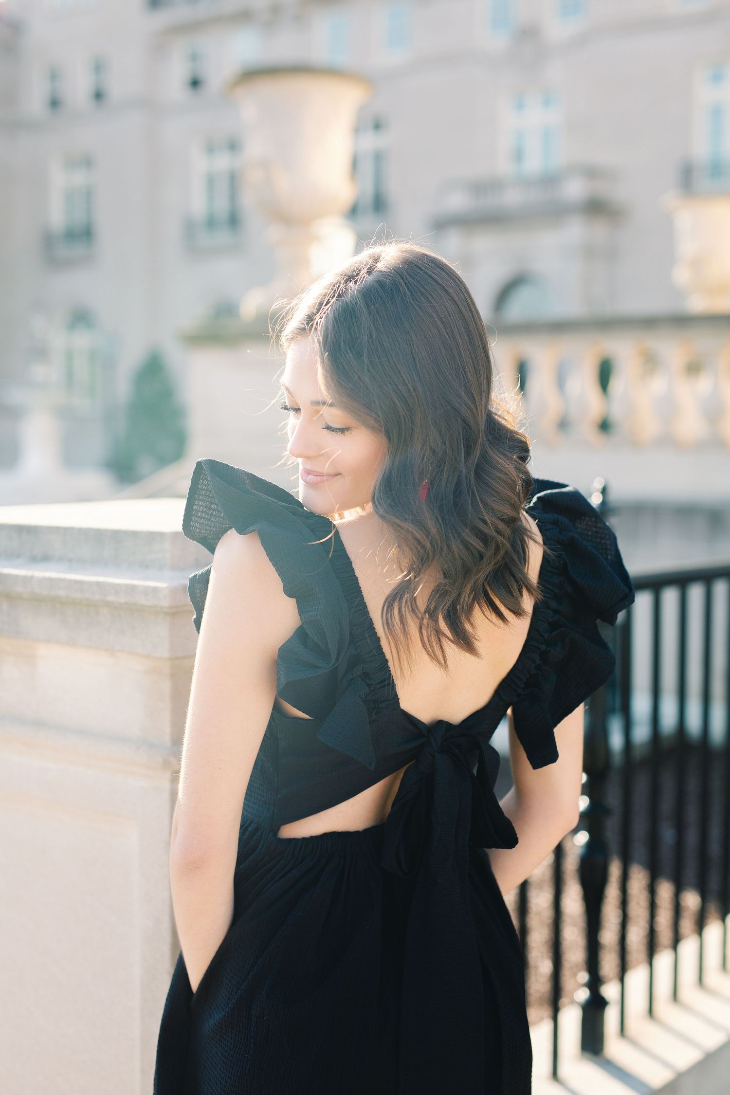 A woman in an elegant black dress with ruffled details poses against a grand architectural backdrop.