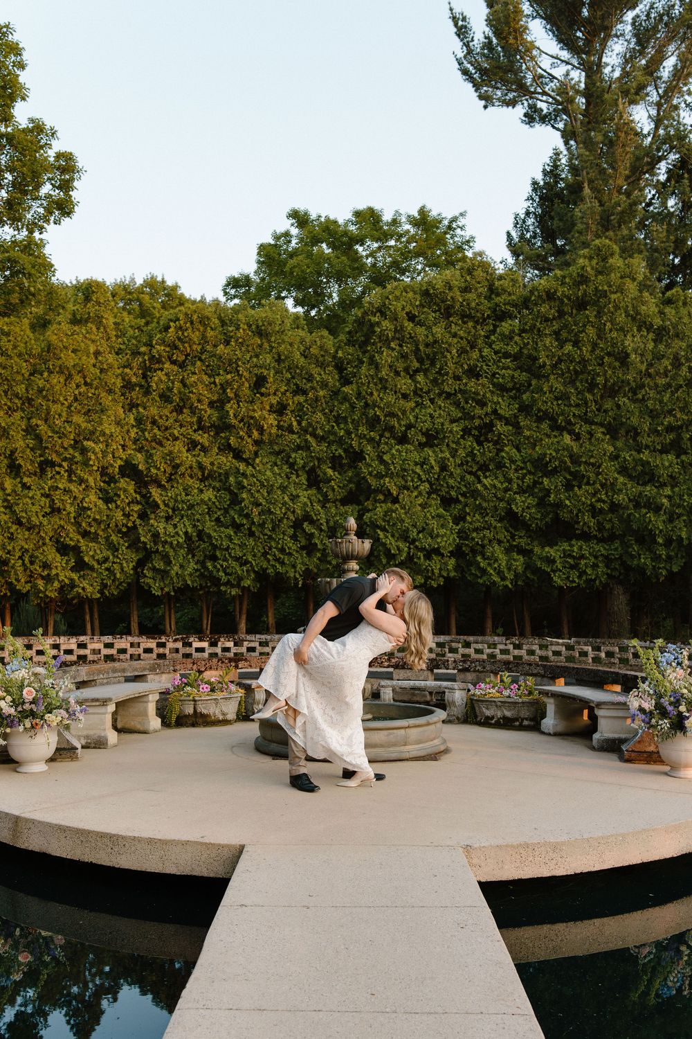 A romantic dip pose on a circular concrete platform surrounded by greenery and water features at sunset.