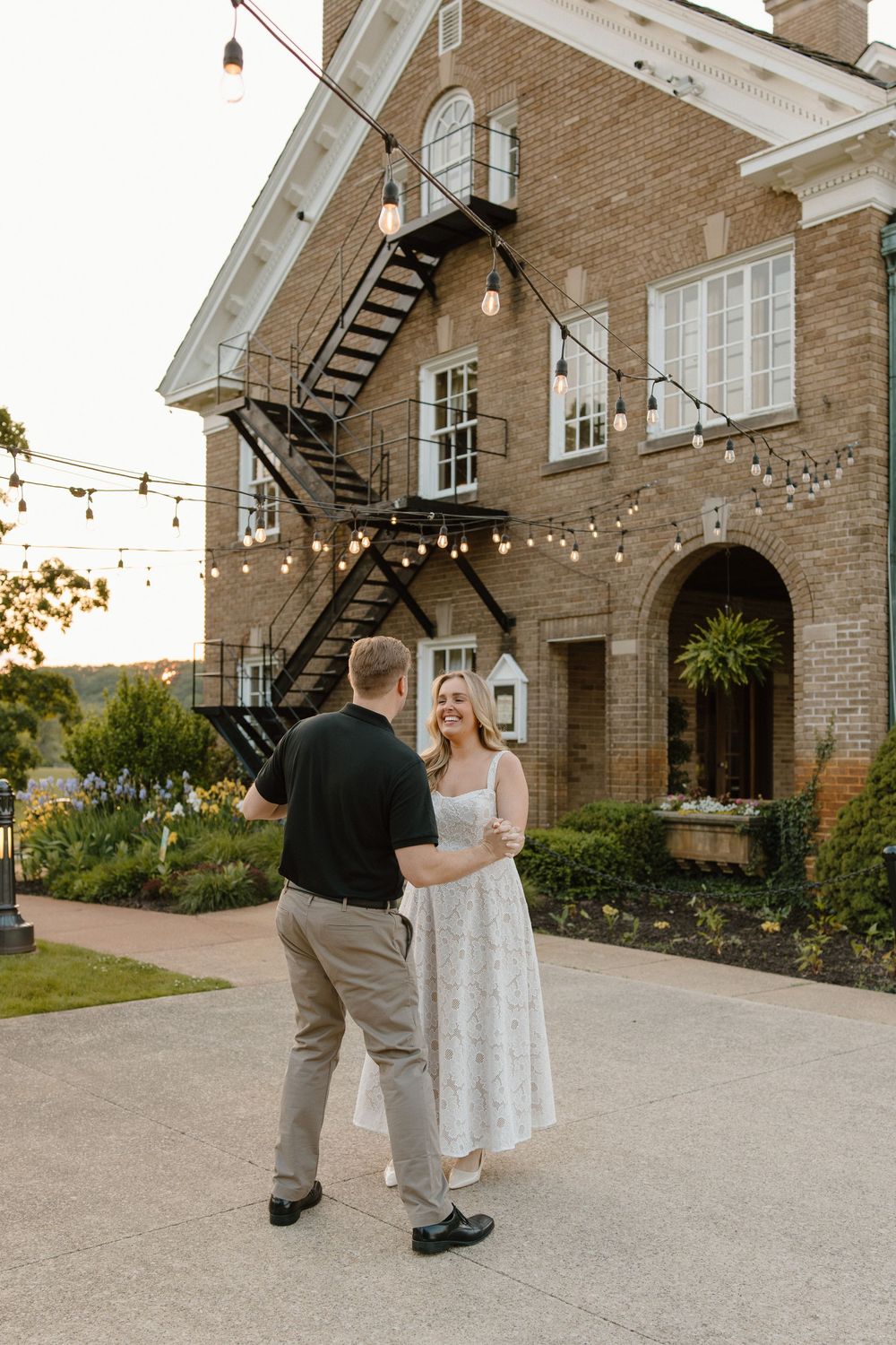 A couple dances outside a brick mansion with twinkling lights and a fire escape.