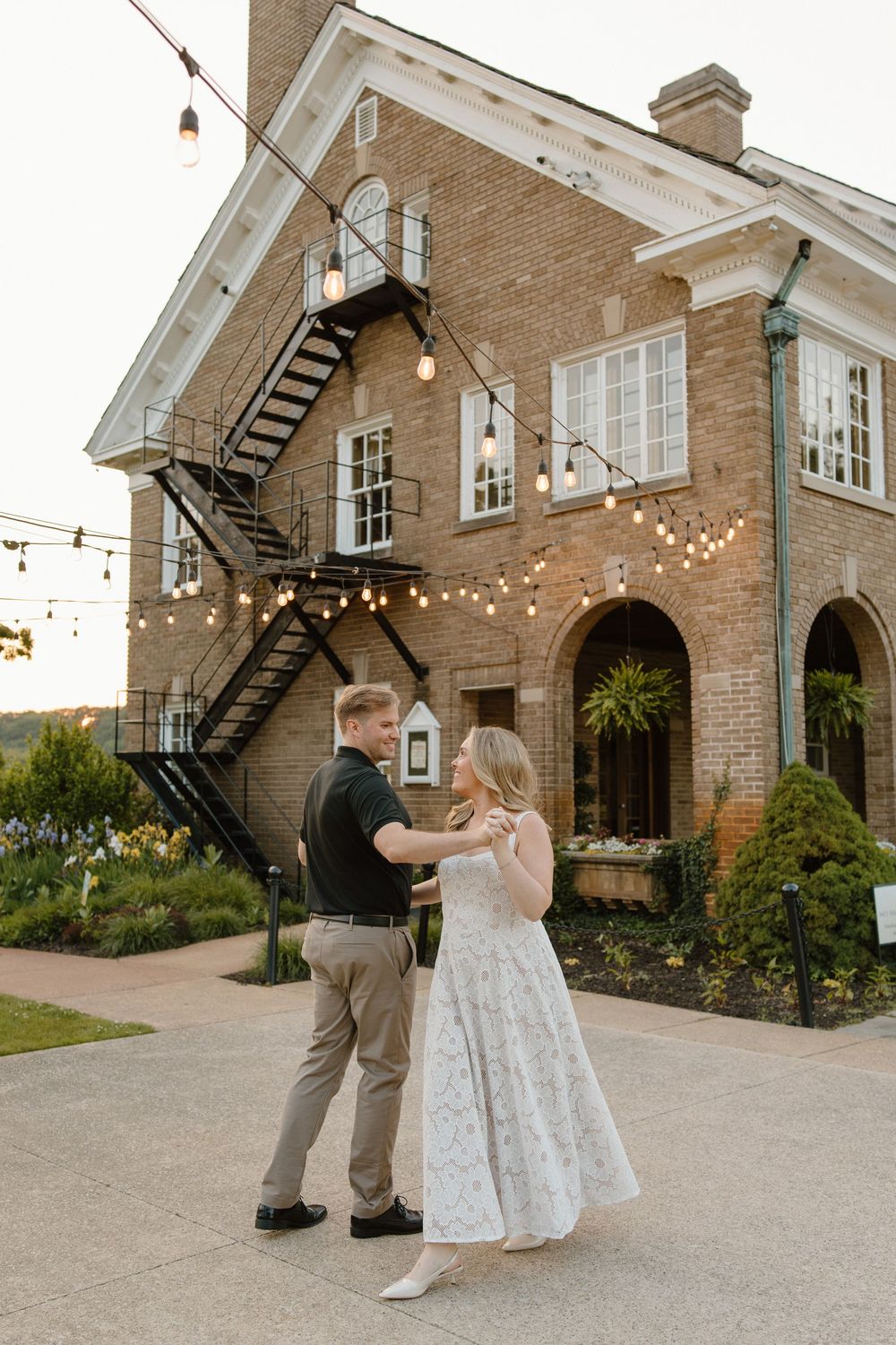 A couple dances together outside a charming brick building decorated with string lights and featuring an outdoor staircase.