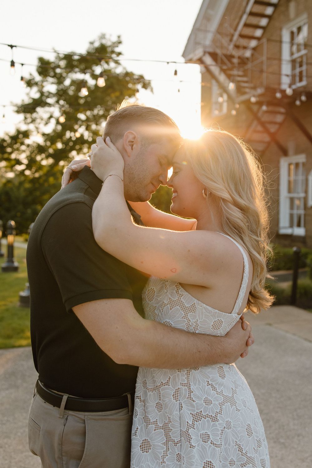 A romantic couple embraces in a golden sunset moment outside a brick building with string lights overhead.
