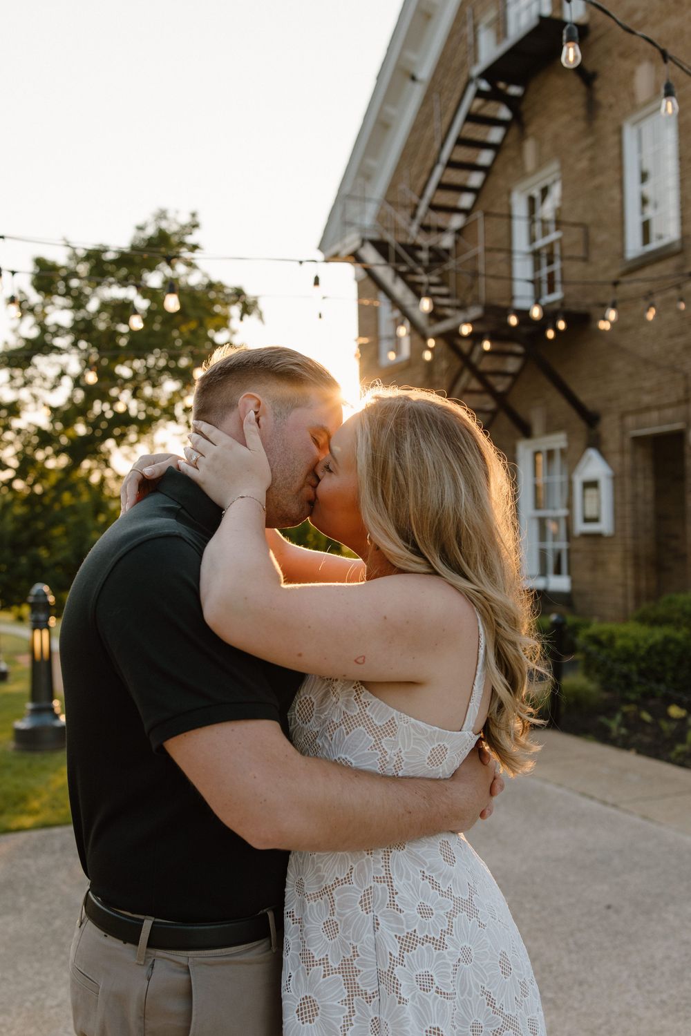 A couple shares a romantic kiss at golden hour outside a brick building with string lights.