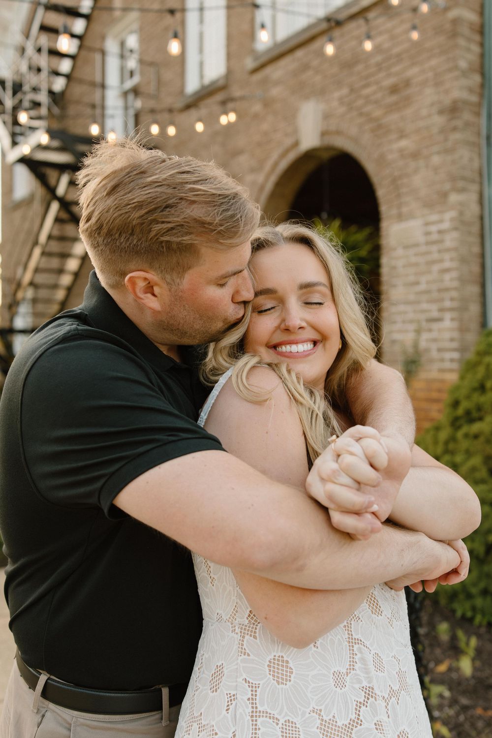 A romantic outdoor photoshoot capturing an intimate embrace against a brick building with string lights.