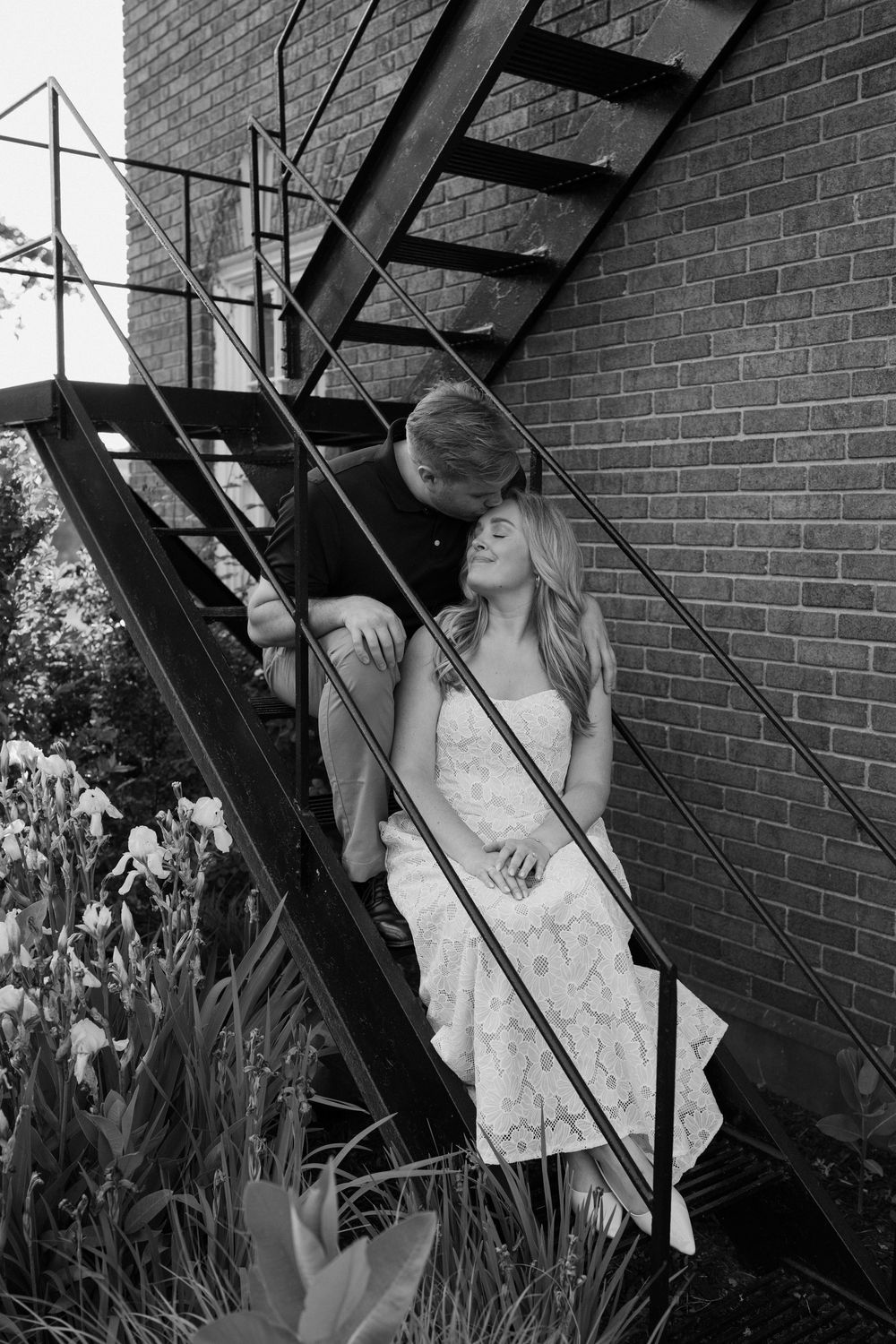 A couple shares an intimate black and white moment on industrial metal stairs against a brick wall backdrop.