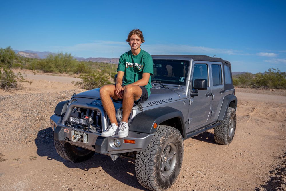 A person in green shirt and shorts sits on the hood of a grey Jeep Wrangler in the desert.