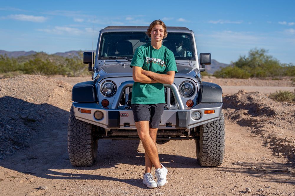 A person in casual wear poses in front of a modified grey Jeep in a desert setting.