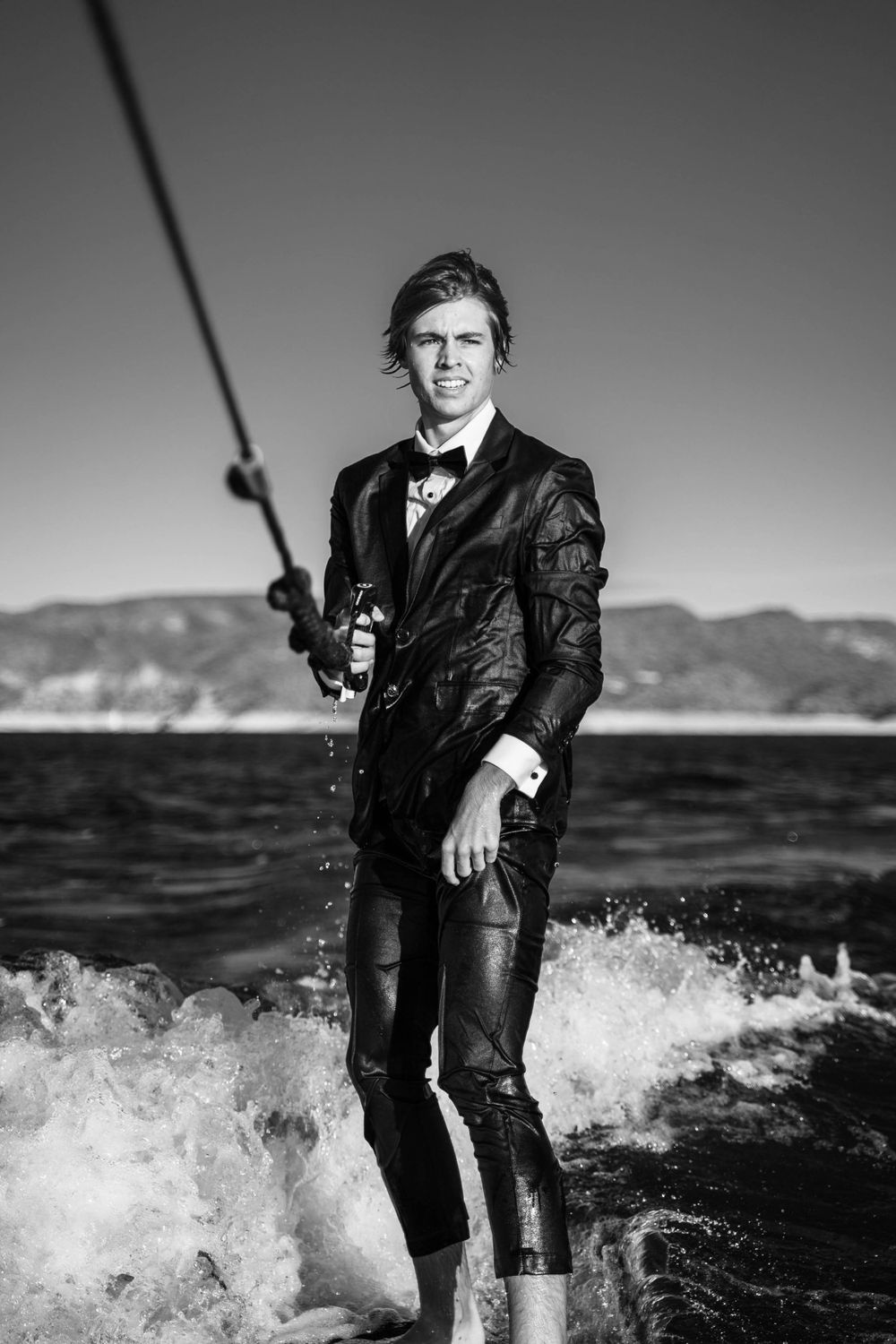 A figure in a formal black tuxedo surfs through ocean waves in a dramatic black and white photo.