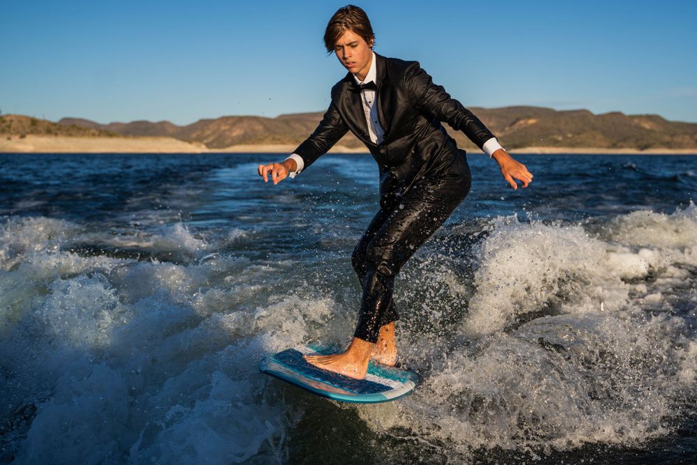 A person in formal attire rides a surfboard through splashing ocean waves near mountains.