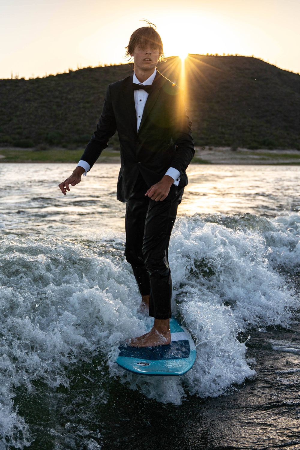 A person wakesurfing in formal wear with a bow tie as the sun sets behind mountains.