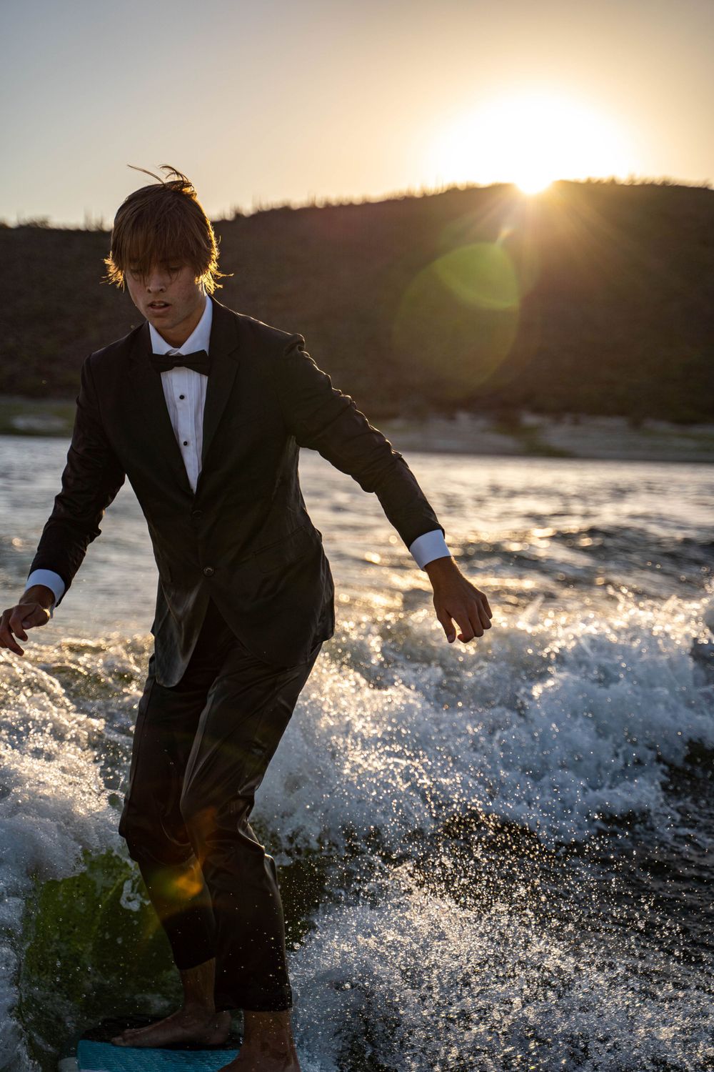 A surfer in formal attire rides waves at sunset with mountains in the background.