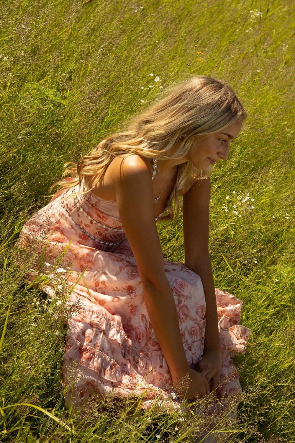 A girl in a pink floral dress sits in a flower field meadow during her senior photo sessions, in Davison Michigan.