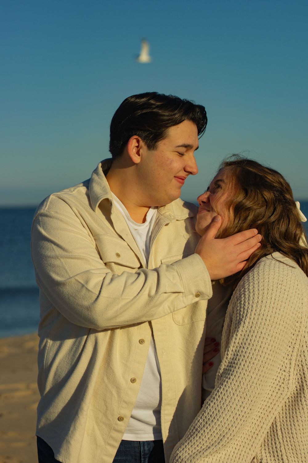 A romantic couple shares an intimate moment on the beach at sunset, with one wearing a beige jacket and the other in a sweater.