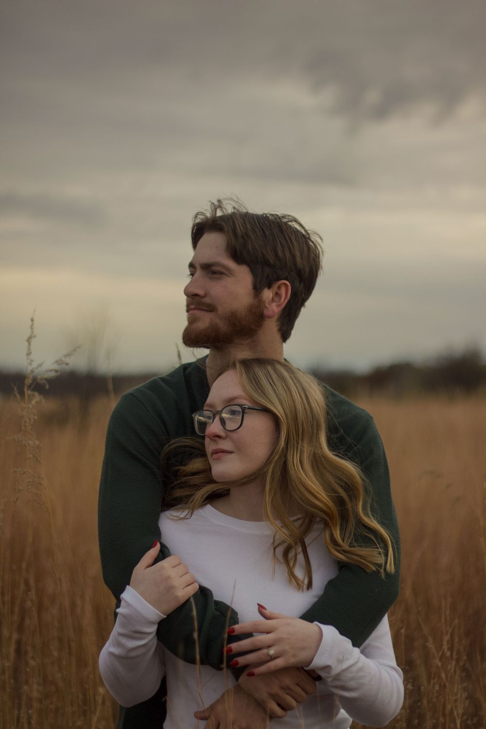 A couple embraces tenderly in a golden wheat field during sunset with moody overcast skies during engagement photos.