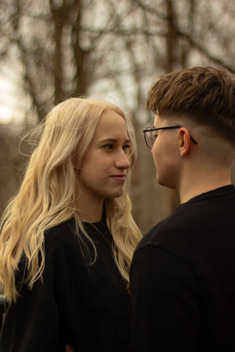 An LGBTQ couple looks into each others eyes surrounded by autumn trees with golden leaves in a park setting during their engagement.