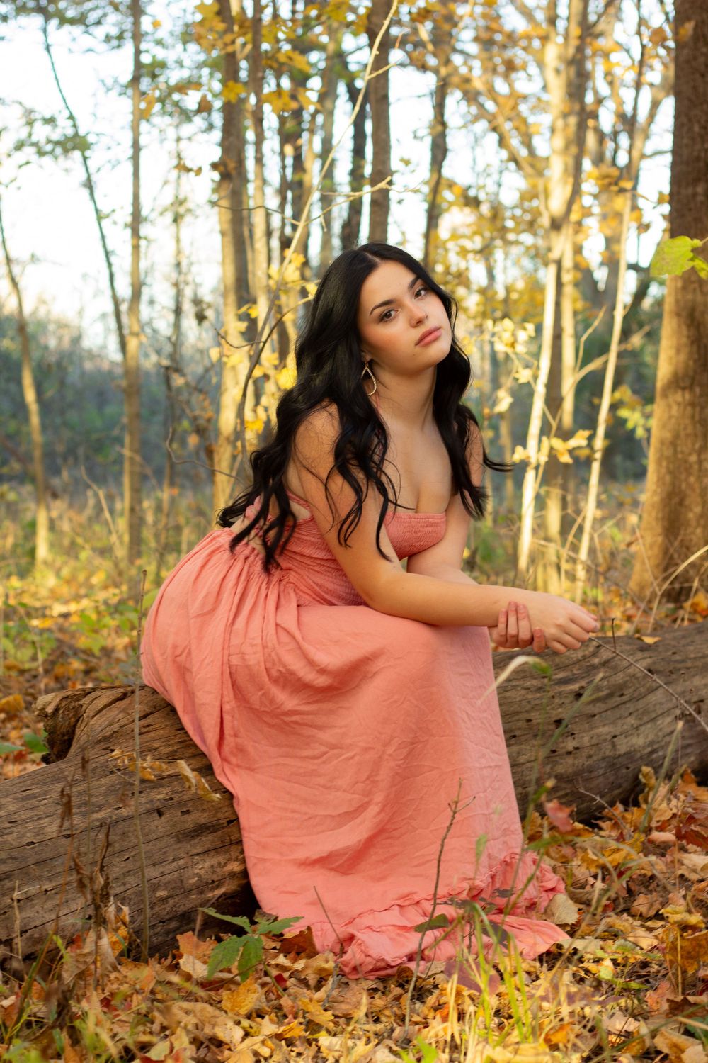 A woman in a coral dress poses gracefully among autumn leaves and trees in a sunlit forest setting.