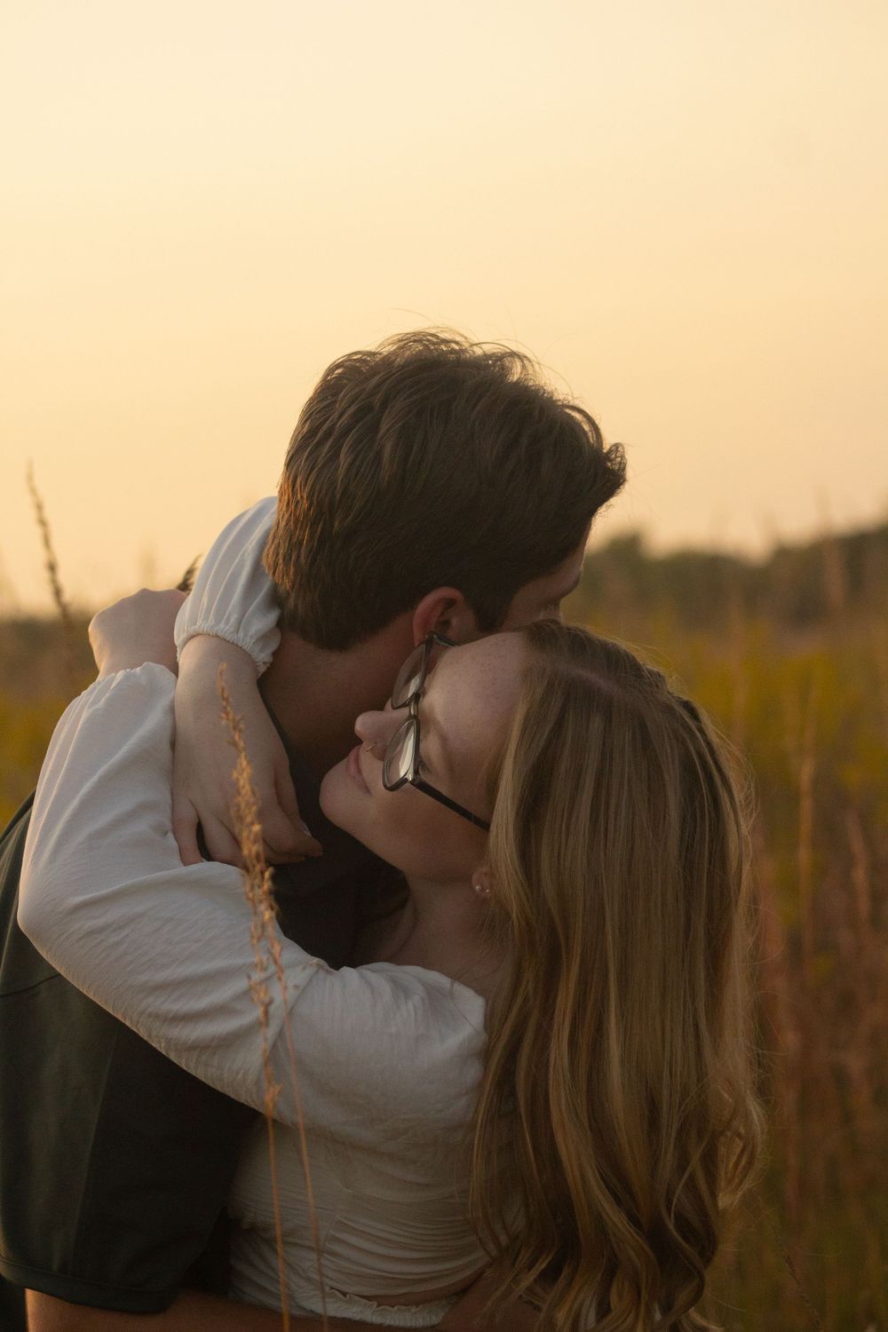 A romantic couple embraces and kisses in a golden wheat field at sunset during engagement photos in Flushing, Michigan.