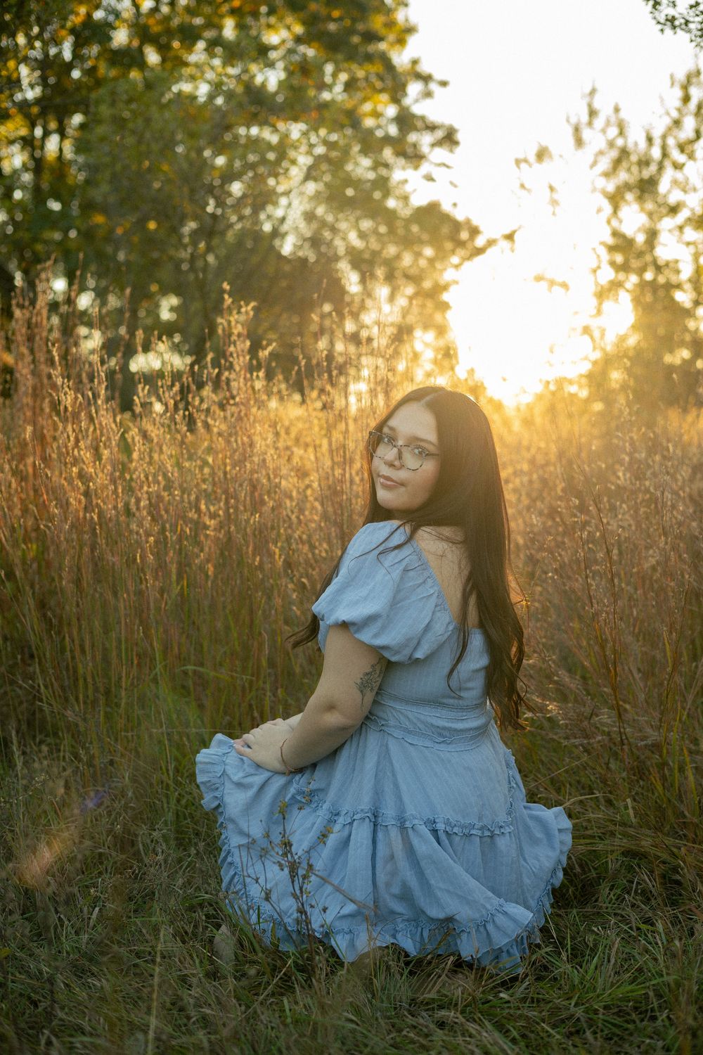 A girl is in a field of tall grass while wearing a blue dress and looks over her shoulder at the camera for her senior photos in Michigan.