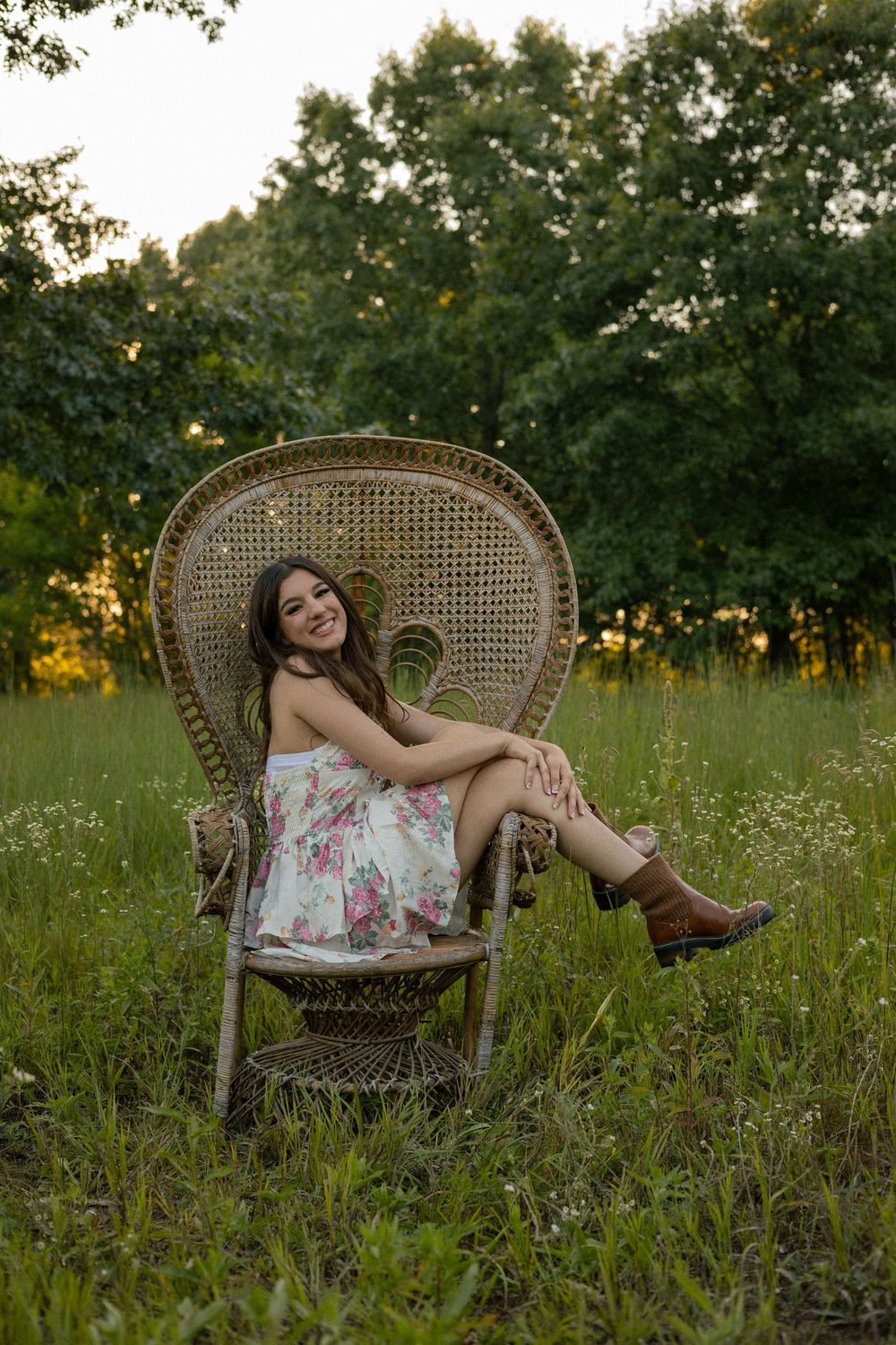 Someone poses casually in a vintage wicker chair surrounded by wildflowers and tall grass during a park senior session in Flushing Michigan.