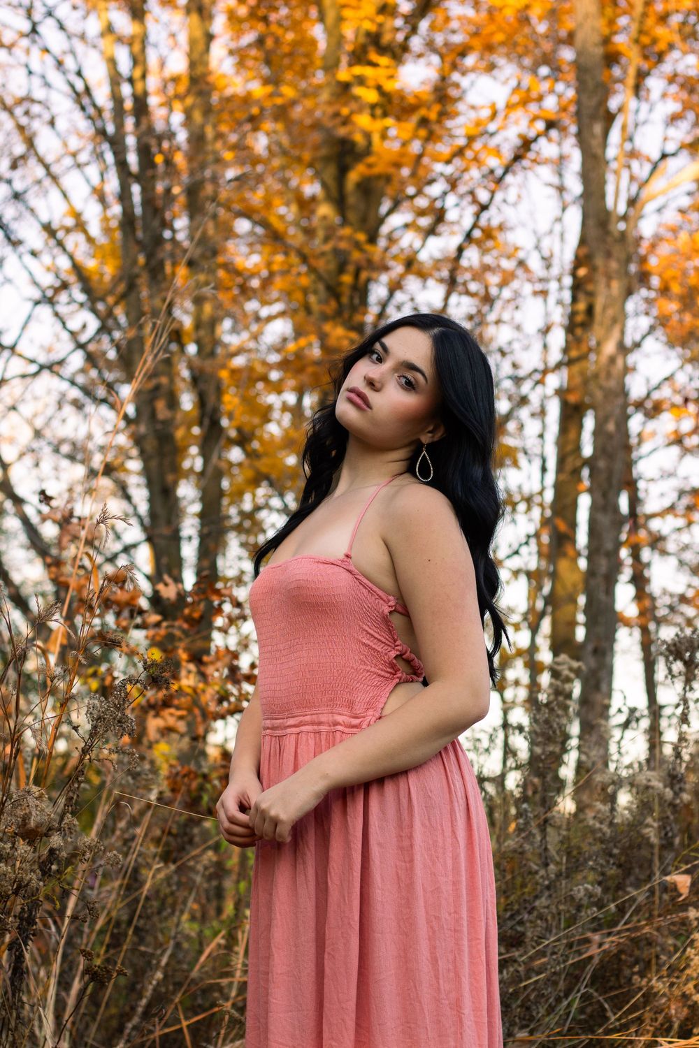 A woman in a coral dress poses gracefully among autumn leaves and trees in a sunlit forest setting.