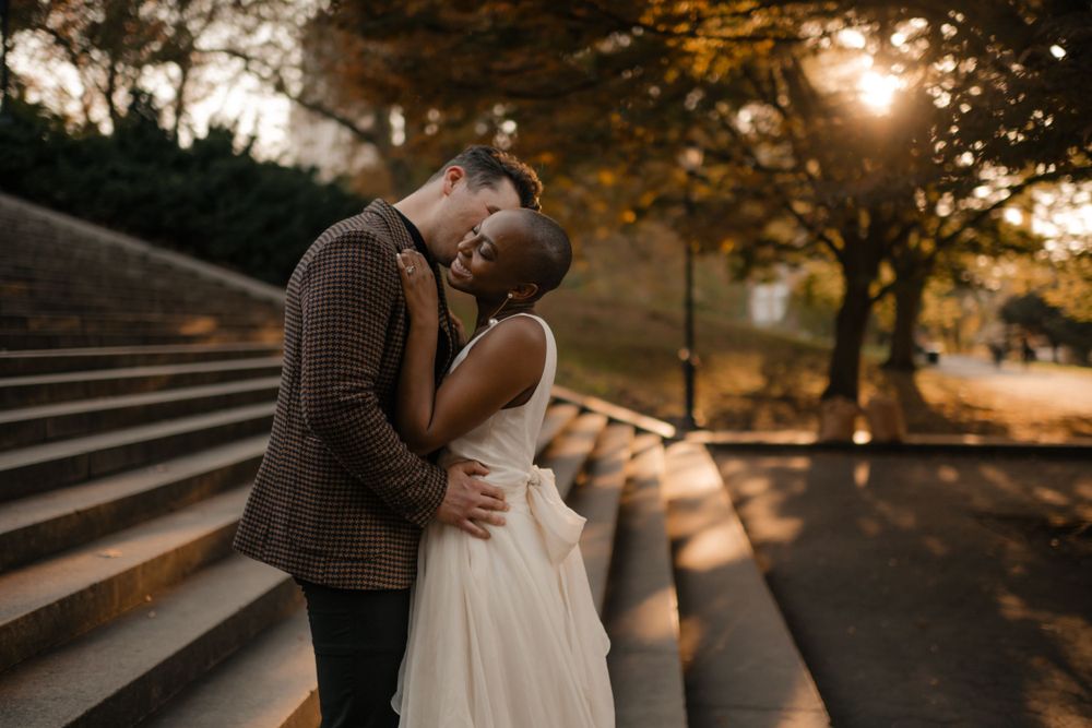 A romantic couple embraces on stone steps at sunset with warm glowing lights and trees in the background.