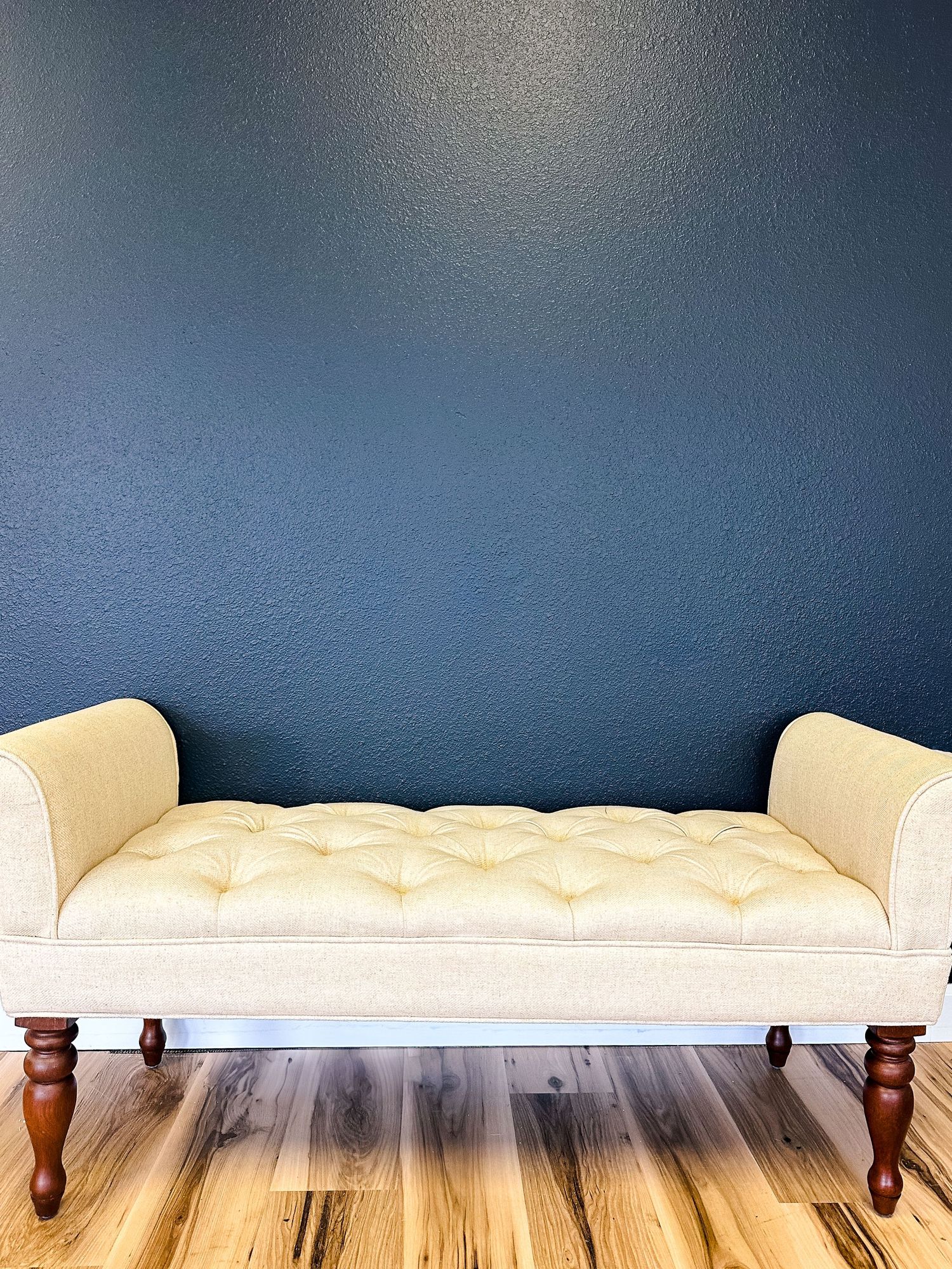 Elegant tufted cream upholstered bench with wooden legs sits against a deep blue wall on hardwood flooring.