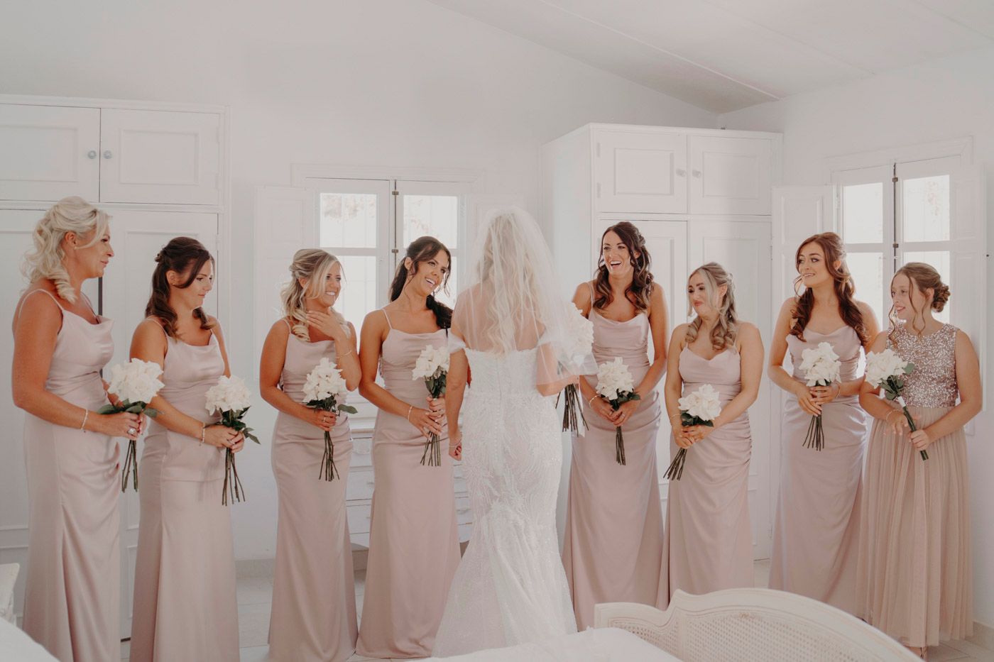 Bridal party in matching blush pink bridesmaid dresses gather in sunlit room with bride in white gown.