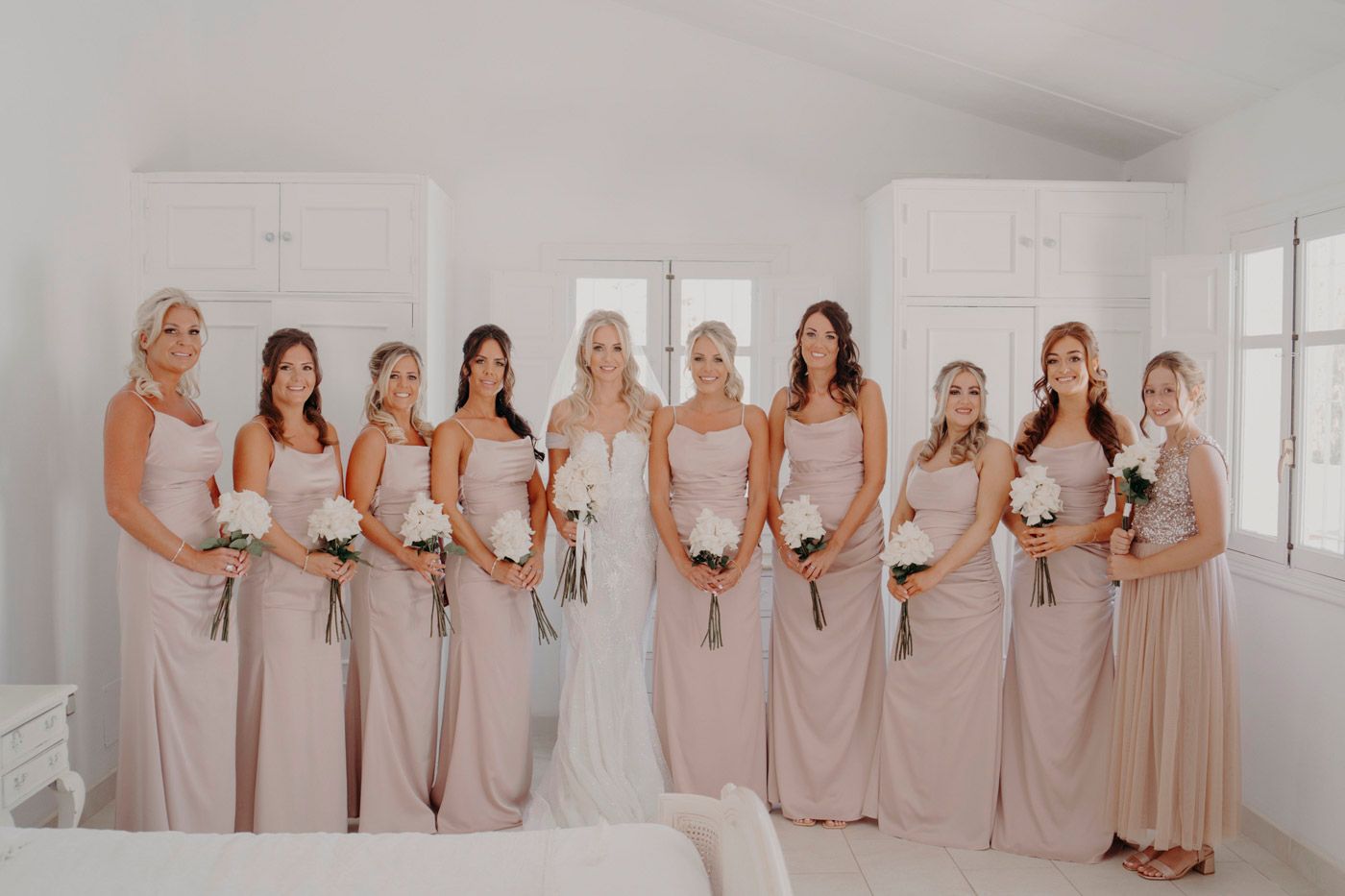 Wedding party of bridesmaids in floor-length blush gowns posing together with bride in bright white dress.