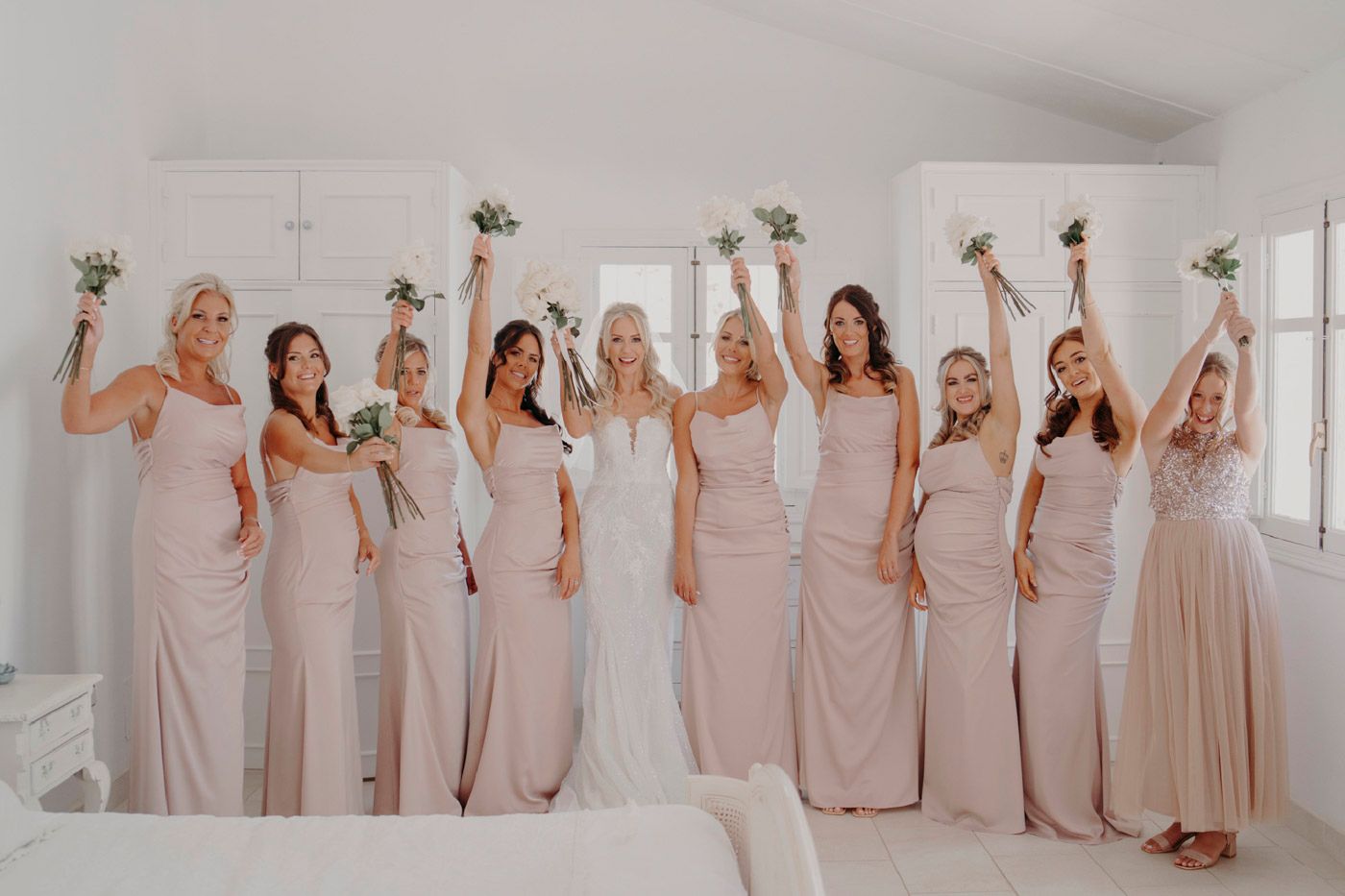 A group of bridesmaids in blush-colored dresses raise their bouquets while celebrating in a bright white room.