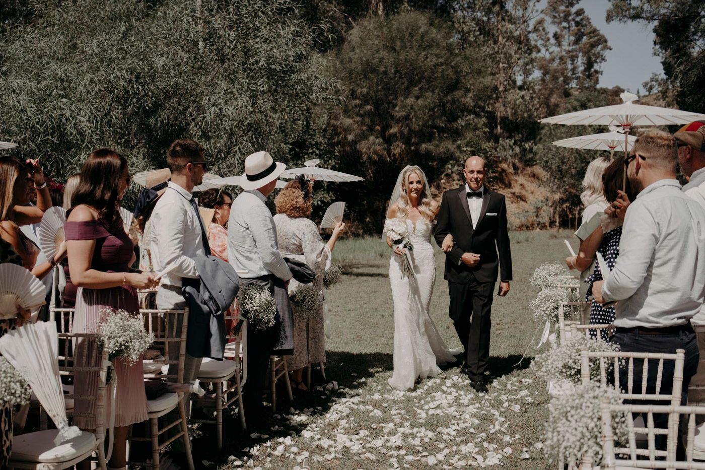 Couple walking down the outdoor wedding aisle surrounded by guests on white chairs and scattered rose petals.