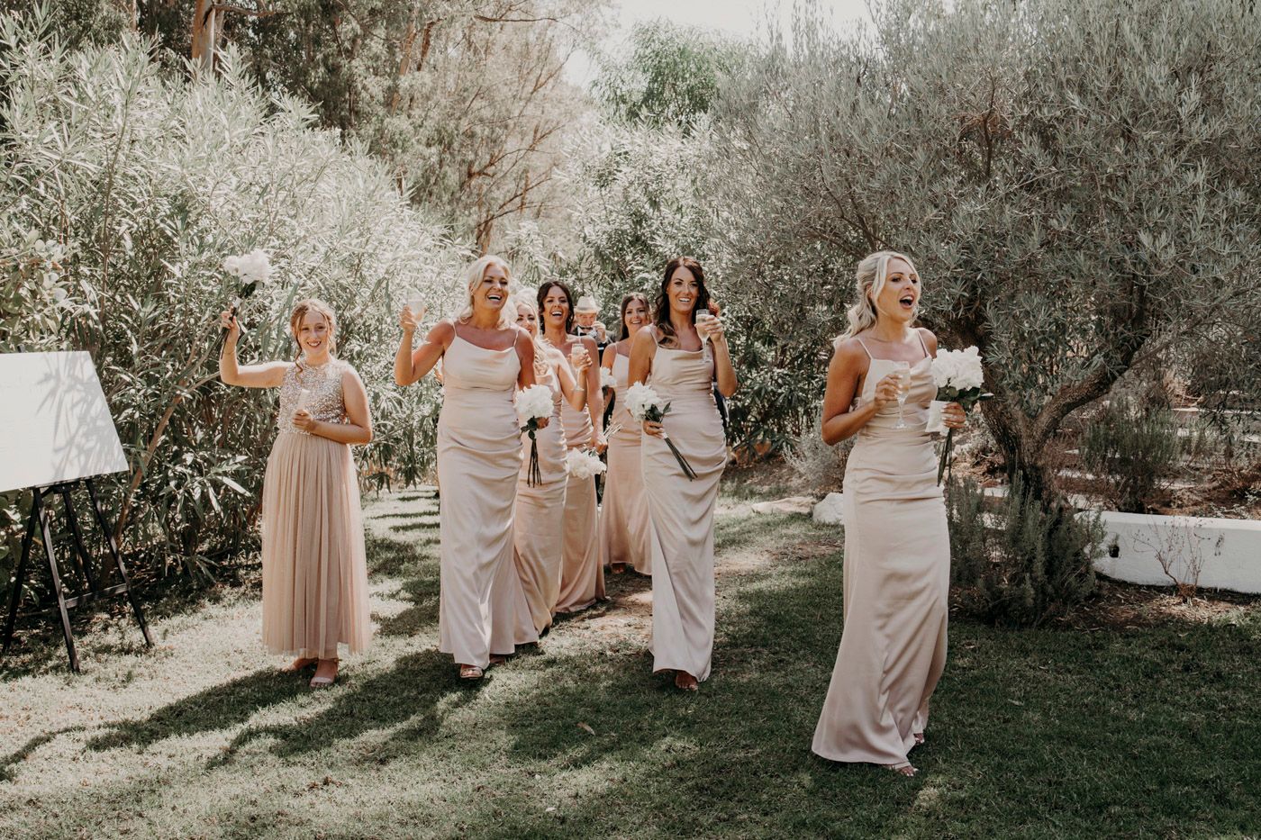 A group of bridesmaids in blush pink dresses walk together through a sunlit garden setting during a wedding celebration.