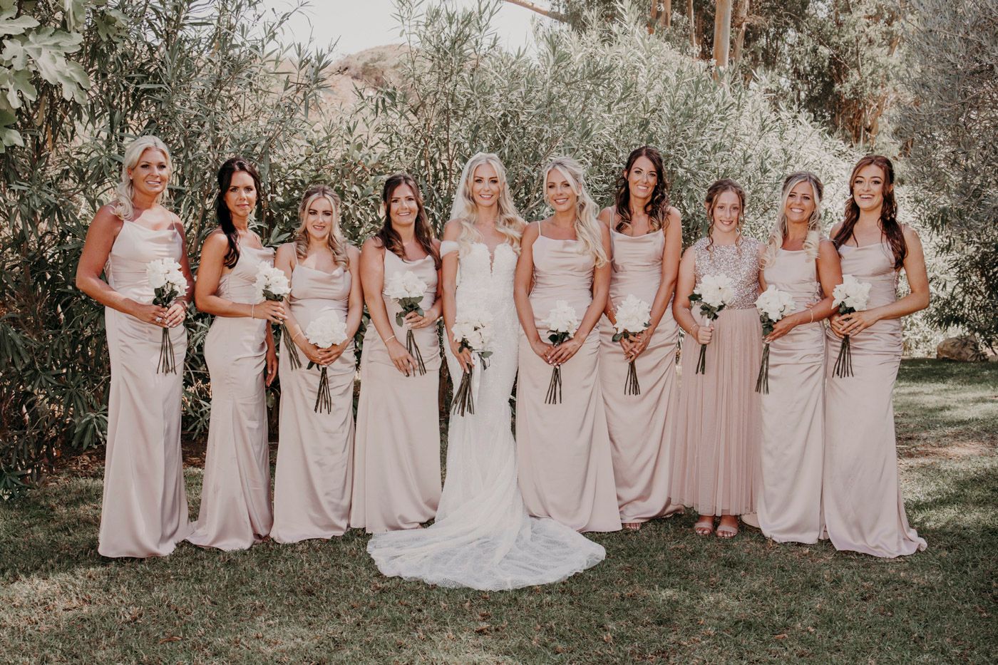A bridal party wearing blush pink bridesmaid dresses poses outdoors among trees on a sunny day.
