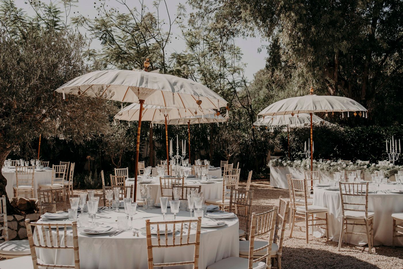 Elegant outdoor wedding reception setup with white parasols, round tables and chiavari chairs in a garden setting.