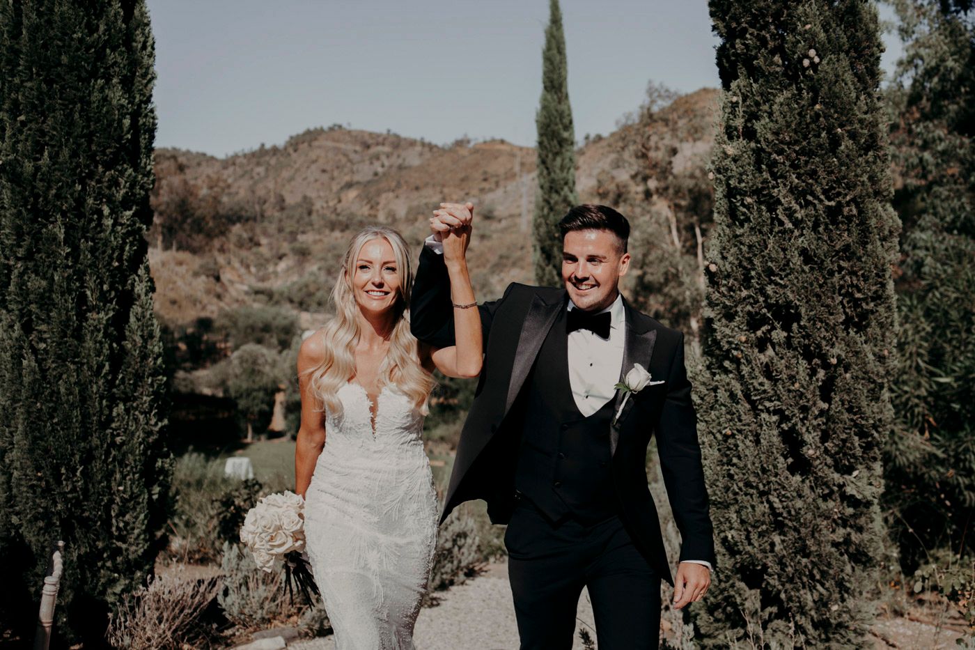 A happy couple walks together on a path lined with tall cypress trees in front of rustic mountain scenery.