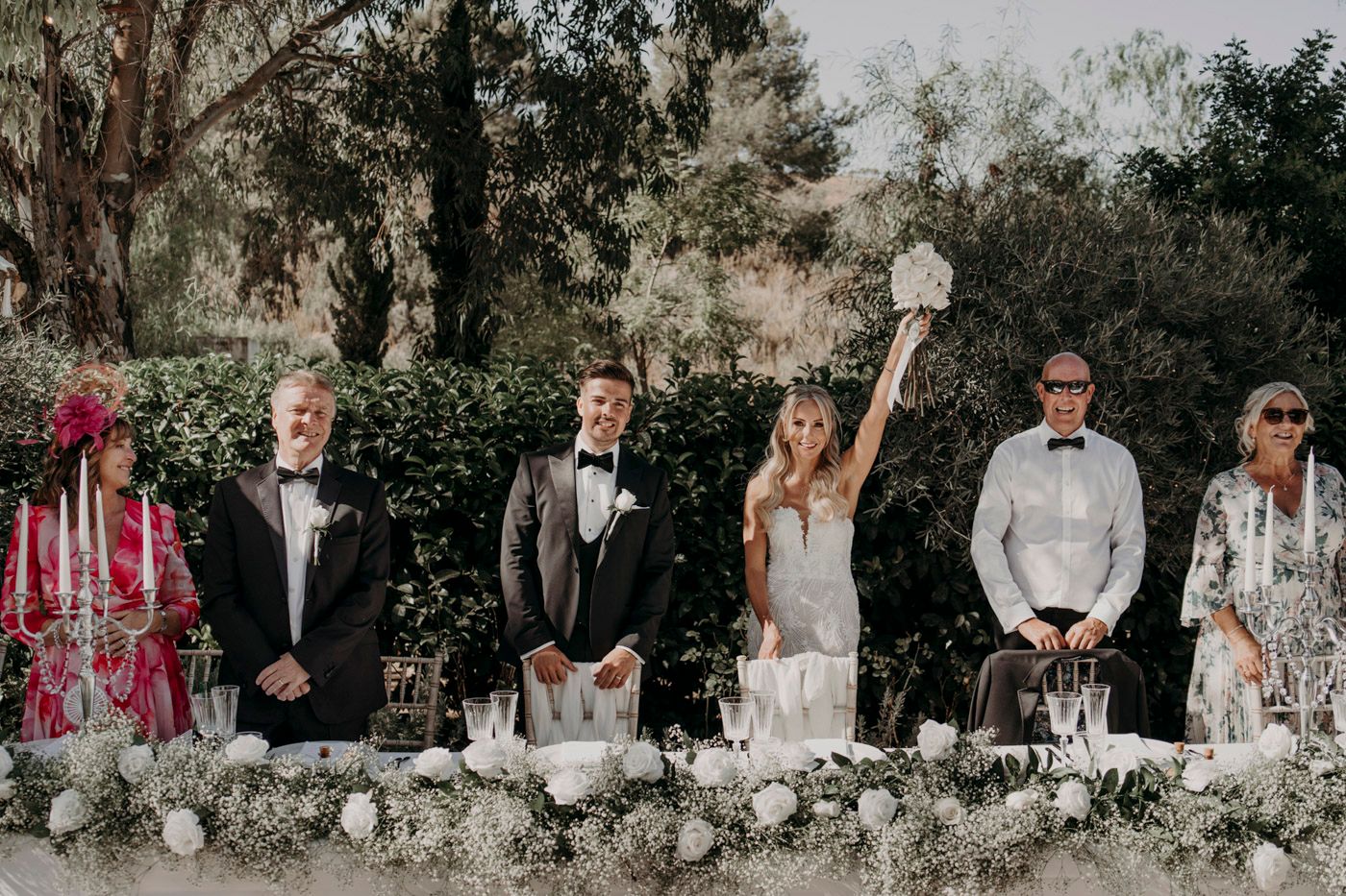 Wedding party stands at a decorated table with pink candles and white floral arrangements in an outdoor garden setting.