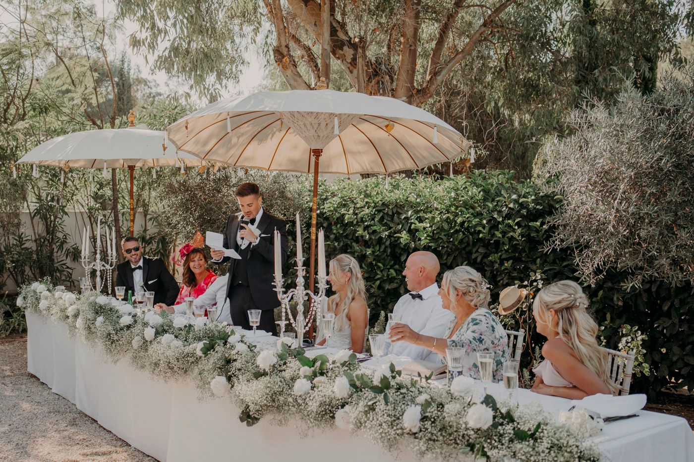 Wedding party sits at an elegant outdoor table decorated with white floral arrangements under a beige umbrella.
