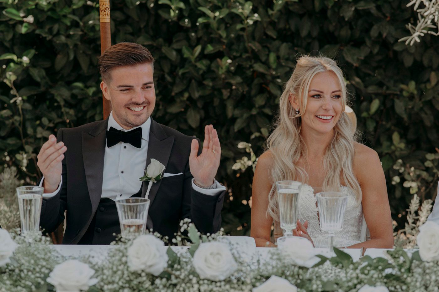 A happy couple sits at their wedding reception table decorated with white flowers and champagne glasses.