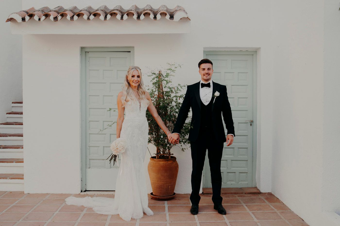 A couple in wedding attire holds hands while standing in front of sage green doors and a terracotta planter.
