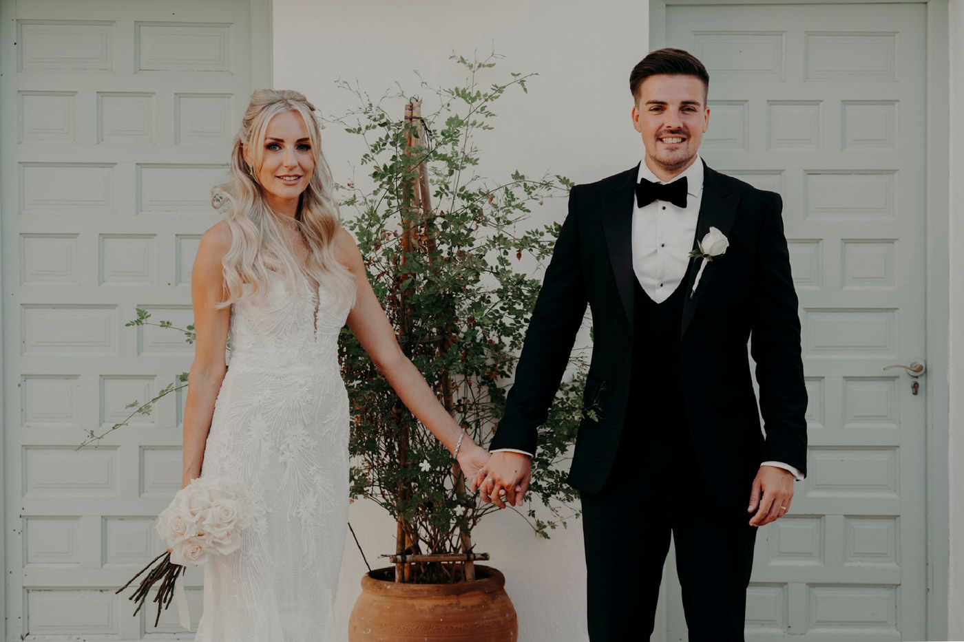A bride and groom hold hands in front of a white garage door with a potted plant decoration.