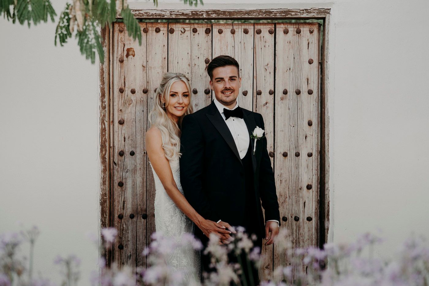 Elegant couple poses in front of rustic wooden door with purple lavender flowers in foreground.