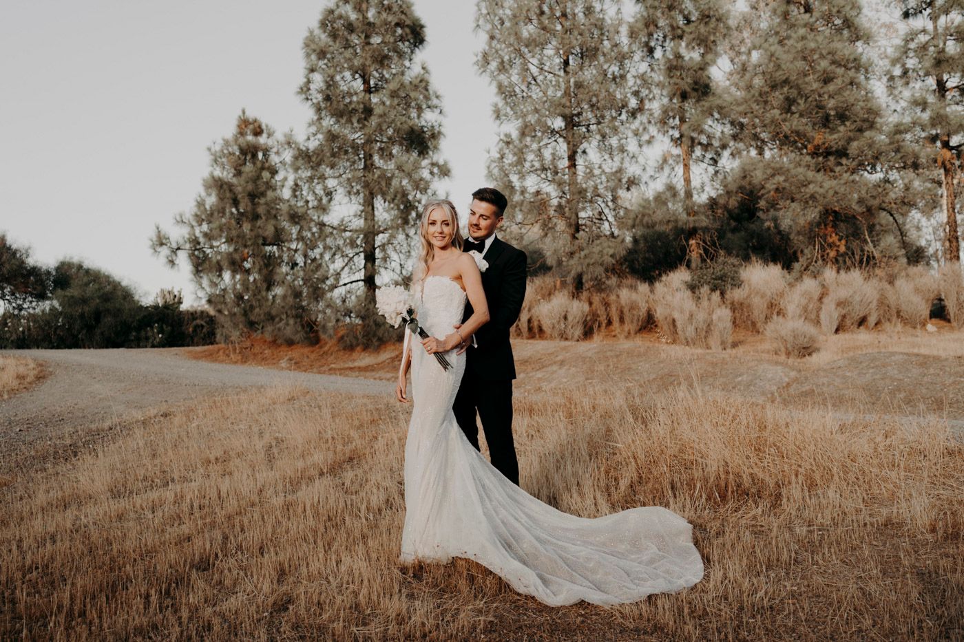 A couple embraces during their wedding photoshoot in a golden field with tall trees at sunset.