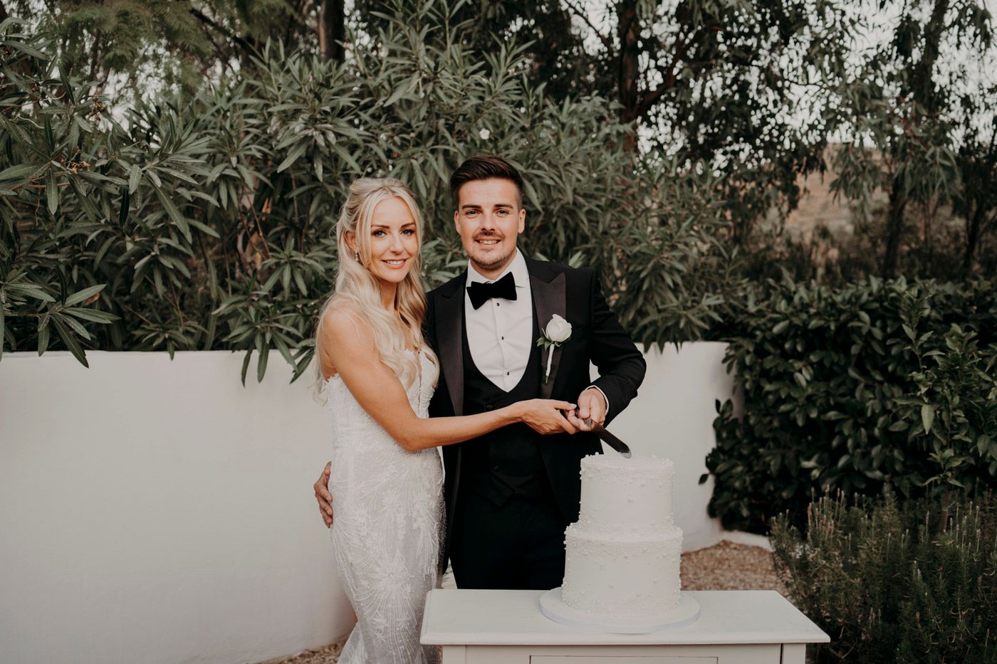 A bride and groom pose together outdoors against a white wall with greenery, dressed in formal wedding attire.