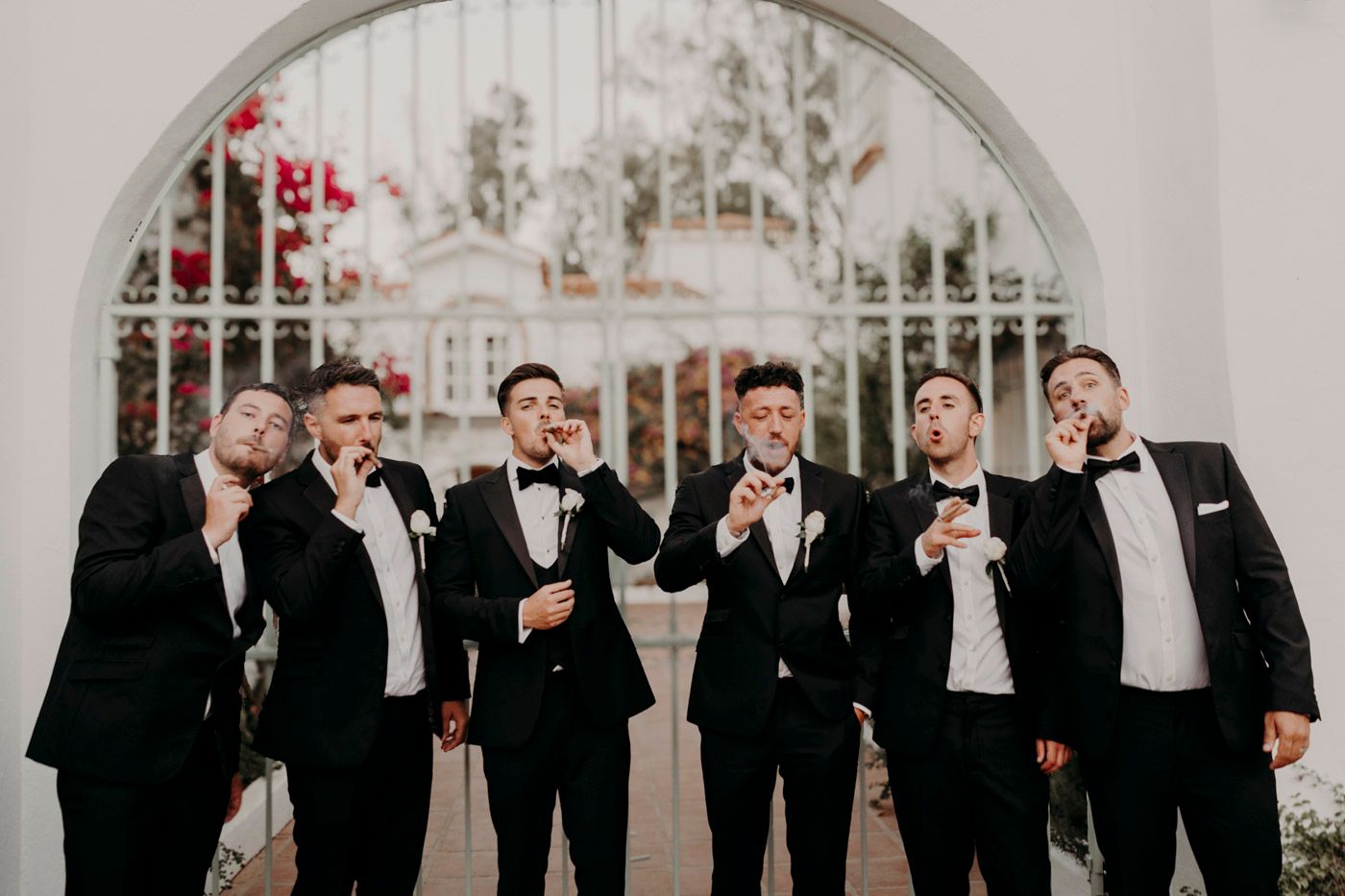 A group of groomsmen in black tuxedos pose playfully under an ornate white archway.