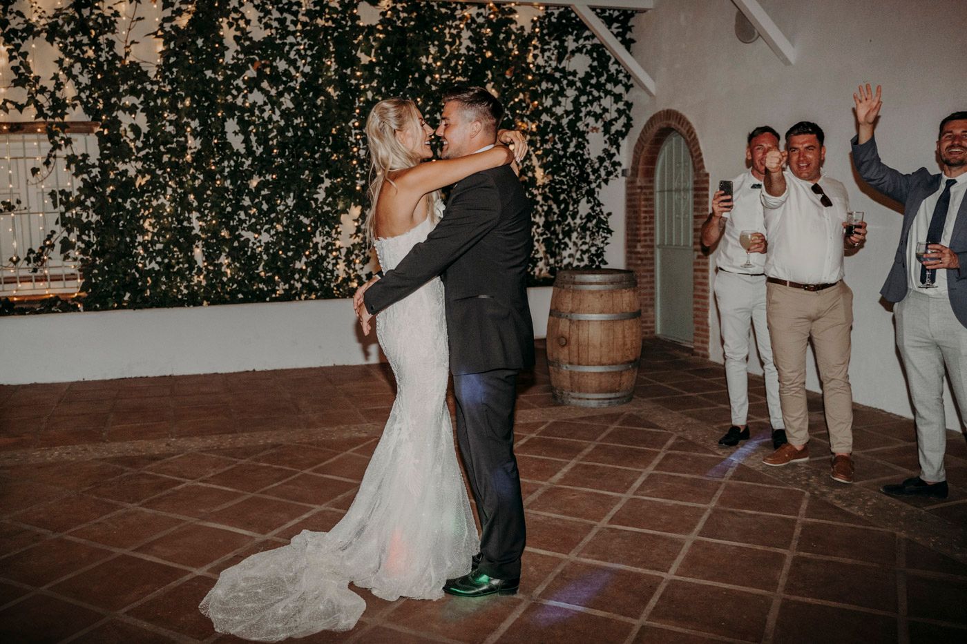 Newlyweds share their first dance on terracotta tiles while guests watch at a rustic wedding reception.
