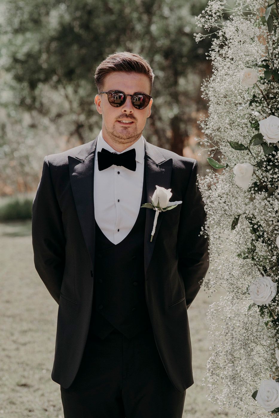A groom in a classic black tuxedo, bowtie and sunglasses stands by white floral wedding decorations outdoors.