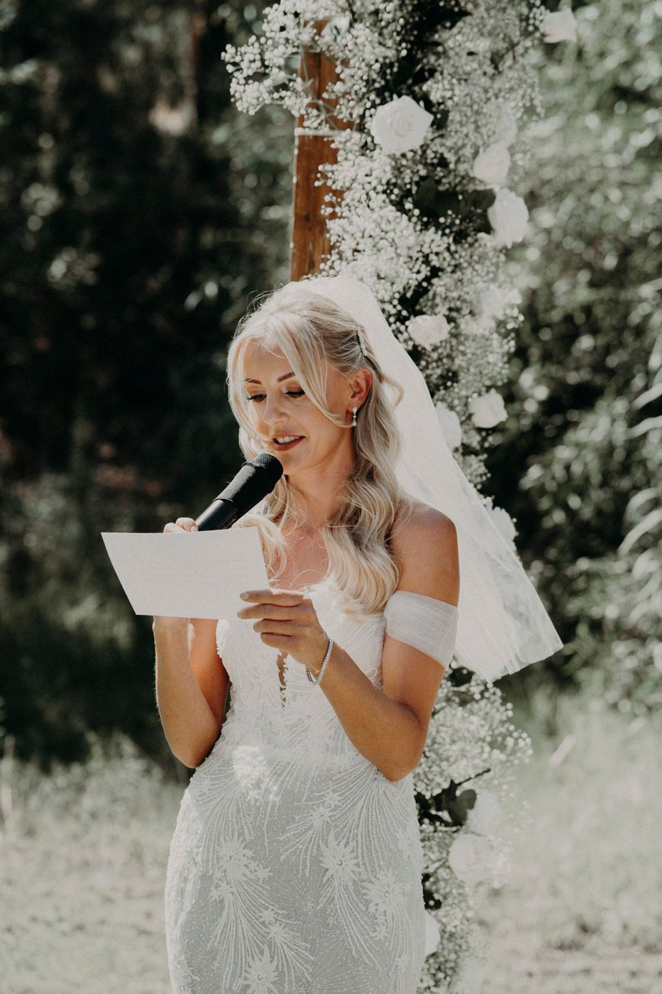 A bride in a detailed white off-shoulder wedding dress reads vows beneath flowering branches on a sunny day.
