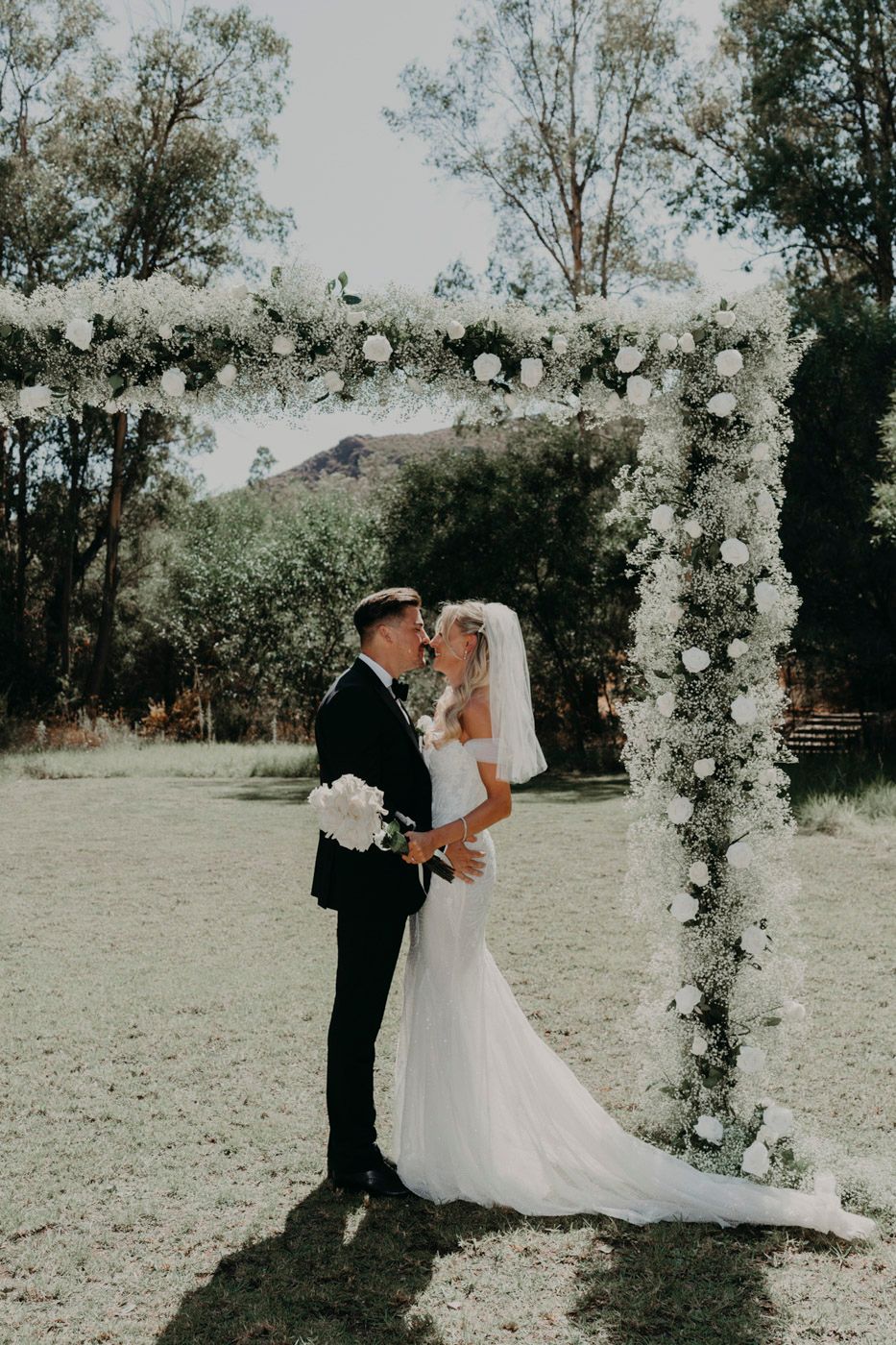 A romantic wedding ceremony under a floral arch adorned with white roses against a natural backdrop.