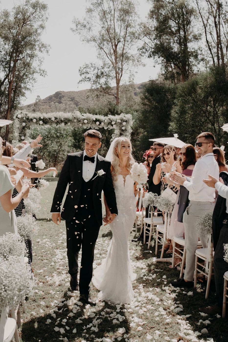Newlyweds walk down the aisle as guests shower them with confetti at an outdoor garden wedding ceremony.